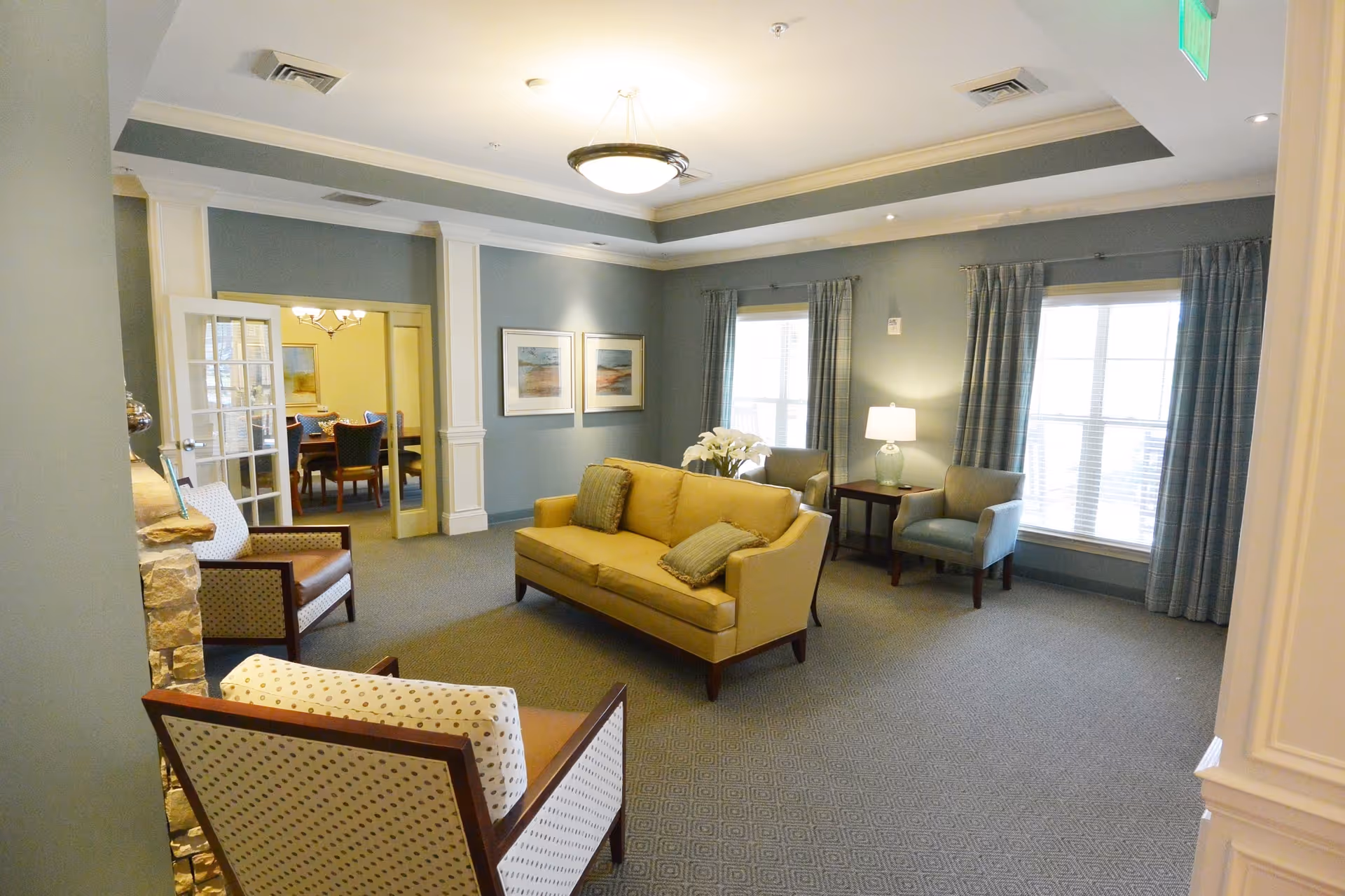A cozy living room area in a senior living facility with a beige sofa, two armchairs, a side table with a lamp, and two large windows with curtains. The walls are painted blue-gray and decorated with framed artwork. In the background, a dining room with a table and chairs is visible through glass double doors.