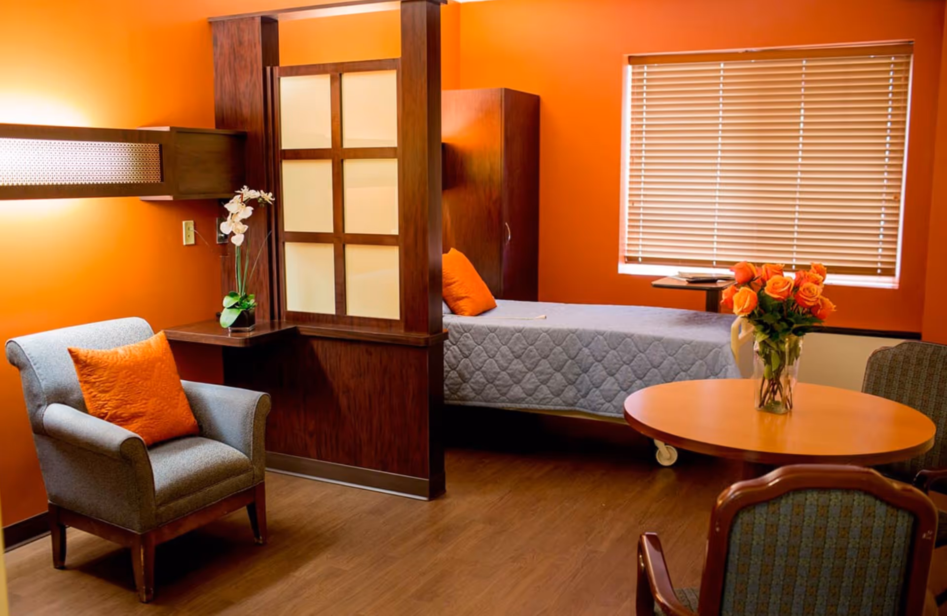 A cozy room with orange walls featuring a single bed with a gray quilt, an armchair with an orange pillow, a round wooden table with a vase of orange roses, and a window with closed blinds. The room has wooden flooring and a partial wooden divider with frosted glass panels.