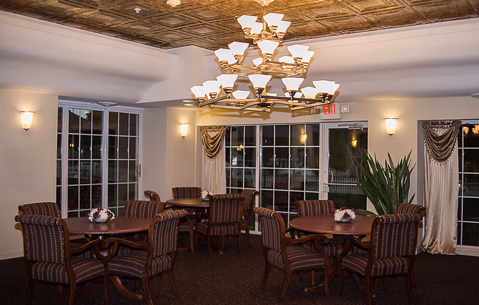 Round dining tables with upholstered chairs beneath a multi-tier chandelier in a communal dining room with sliding glass doors.