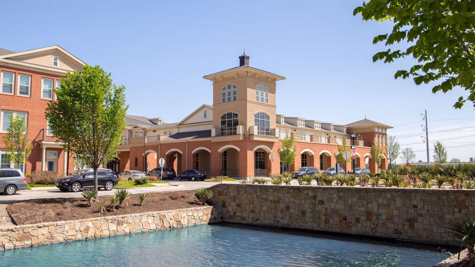 Exterior view of a multi-story assisted living building with arched entrances, parked cars, and a stone-lined pond in the foreground.