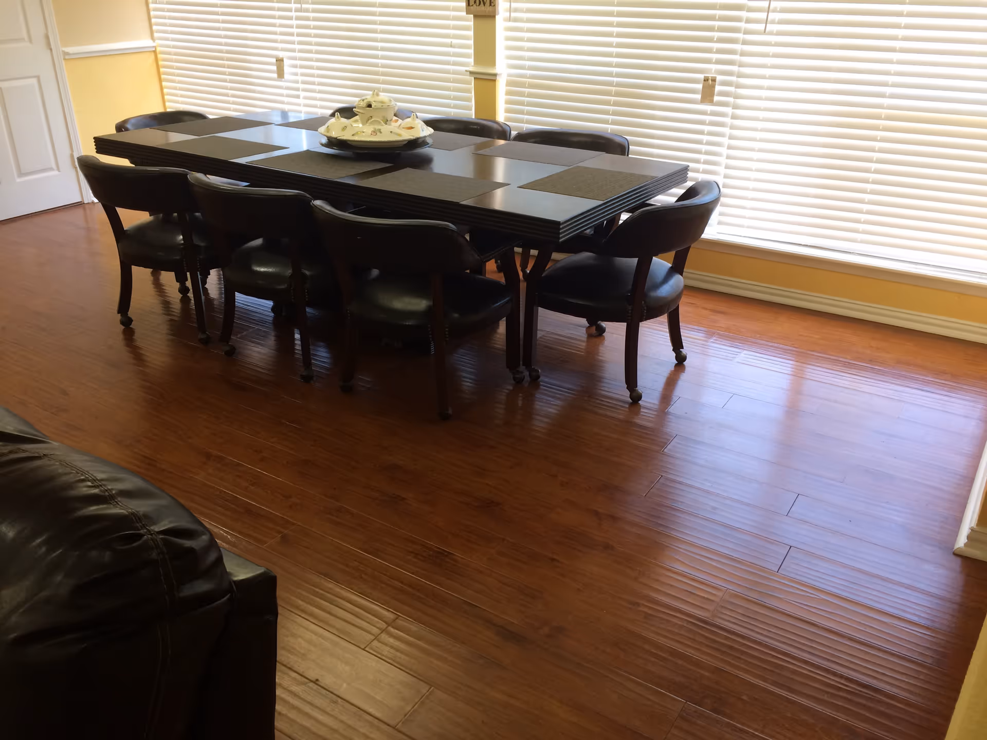A dining room with a dark wooden rectangular table surrounded by eight black cushioned chairs on wheels. The table has black placemats and a decorative white ceramic dish set in the center. The room has wooden flooring and large windows with closed white blinds, allowing natural light to fill the space. A portion of a black leather sofa is visible in the foreground.