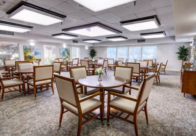 A spacious dining room with multiple round wooden tables and cushioned chairs arranged neatly. The room has large windows allowing natural light to brighten the space, with paintings on the walls and potted plants adding decorative touches. The ceiling features recessed lighting panels.