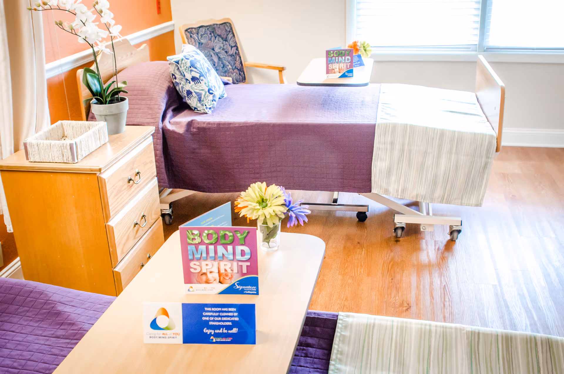 A bright and clean senior living bedroom with two beds covered in purple bedspreads and light striped blankets. A wooden nightstand with a potted orchid and a wicker basket is beside one bed. A small table between the beds holds colorful flowers and a sign that reads 'BODY MIND SPIRIT'. The room has wooden flooring and a window with blinds letting in natural light.