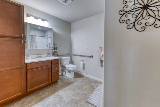 A clean and spacious bathroom featuring a wooden vanity with a large mirror and three light fixtures above it. There is a white toilet with a grab bar next to it, a towel rack with towels, and a decorative metal wall art on the right wall. The floor is tiled and there is a white bath mat near the entrance.