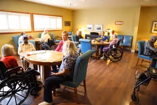 A group of elderly people sitting around tables in a well-lit room with wooden floors and large windows. Some individuals are in wheelchairs, and others are seated in chairs, engaging in conversation and activities. The room has a cozy atmosphere with light-colored walls and framed pictures.