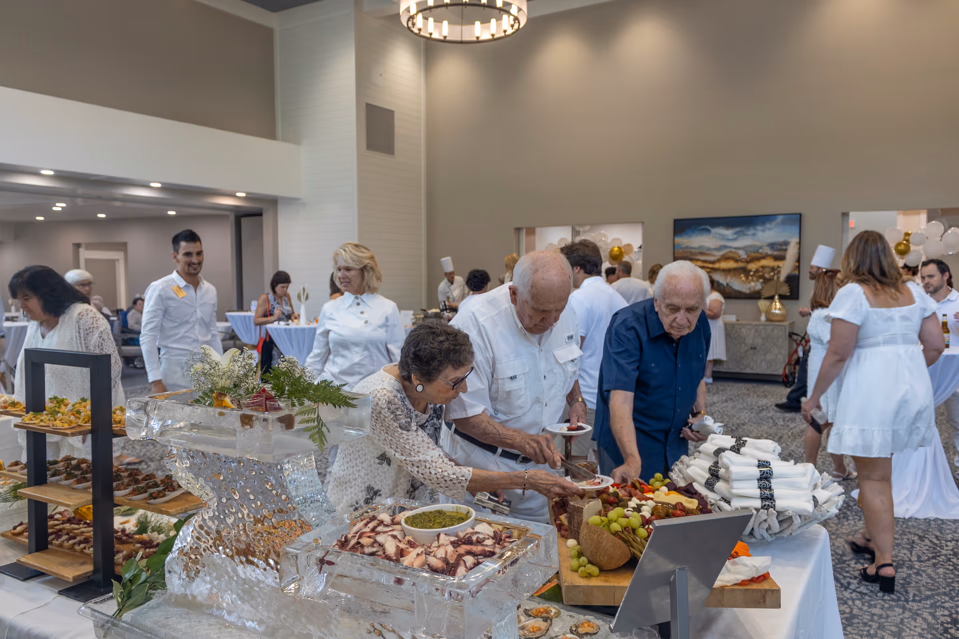 A group of elderly people and staff serving themselves food from a buffet table in a spacious, well-lit room with high ceilings. The buffet includes various appetizers and a fruit platter. People are dressed in casual and semi-formal attire, and some staff members wear white uniforms. The room has modern decor with a large chandelier and artwork on the walls.