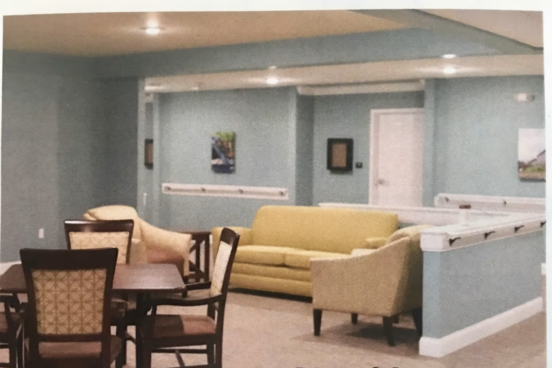 Interior view of a senior living facility common area with a yellow sofa, two beige armchairs, a dark wooden table with four chairs, light blue walls, and framed artwork on the walls.