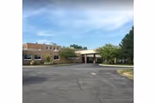 Front exterior of a low-rise brick senior living building with a covered entrance, parking lot, and trees under a blue sky.