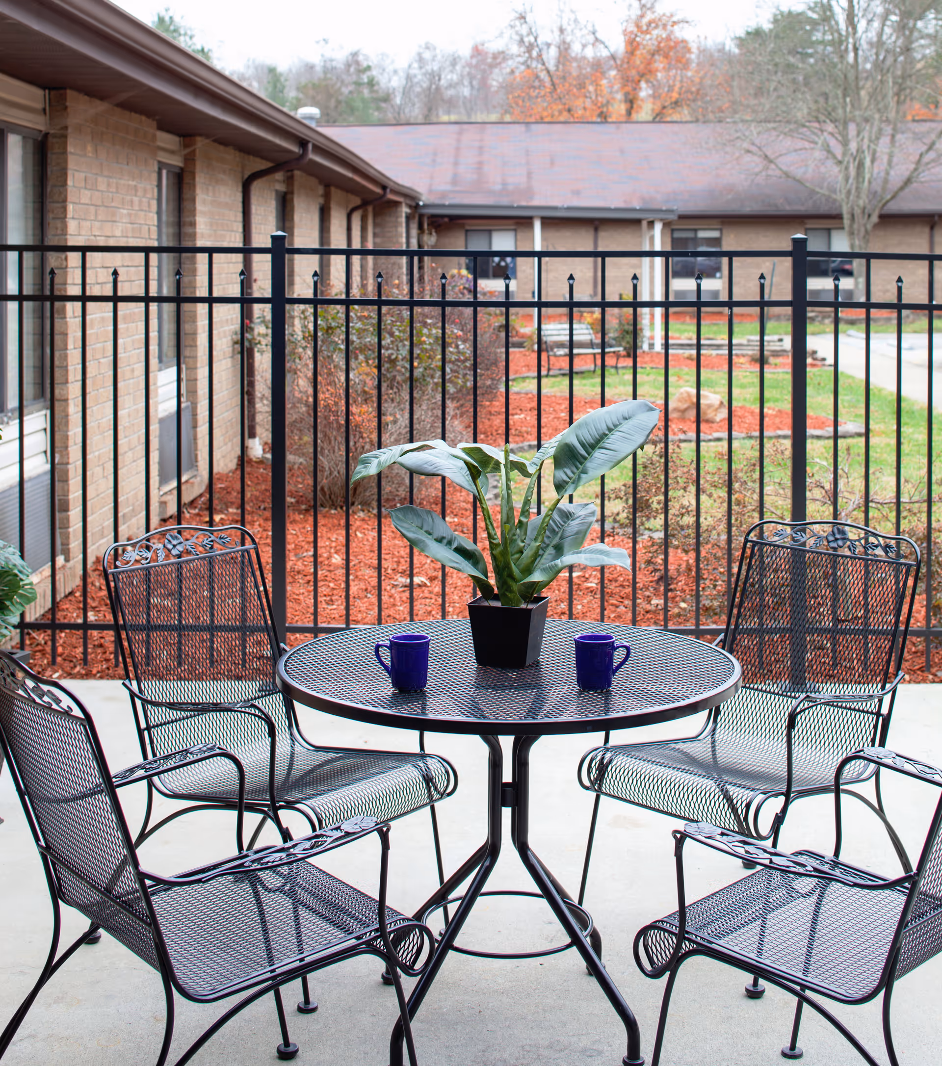A fenced outdoor patio with a round metal table, four matching chairs, a potted plant and two blue mugs in front of a brick building.