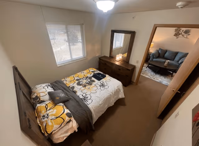 A bedroom with a wooden bed frame and a bed made with white, yellow, and gray floral bedding. There is a wooden dresser with a large mirror on top, and a window with blinds letting in natural light. An open door leads to a living room area with a blue couch, a coffee table, and a lamp.