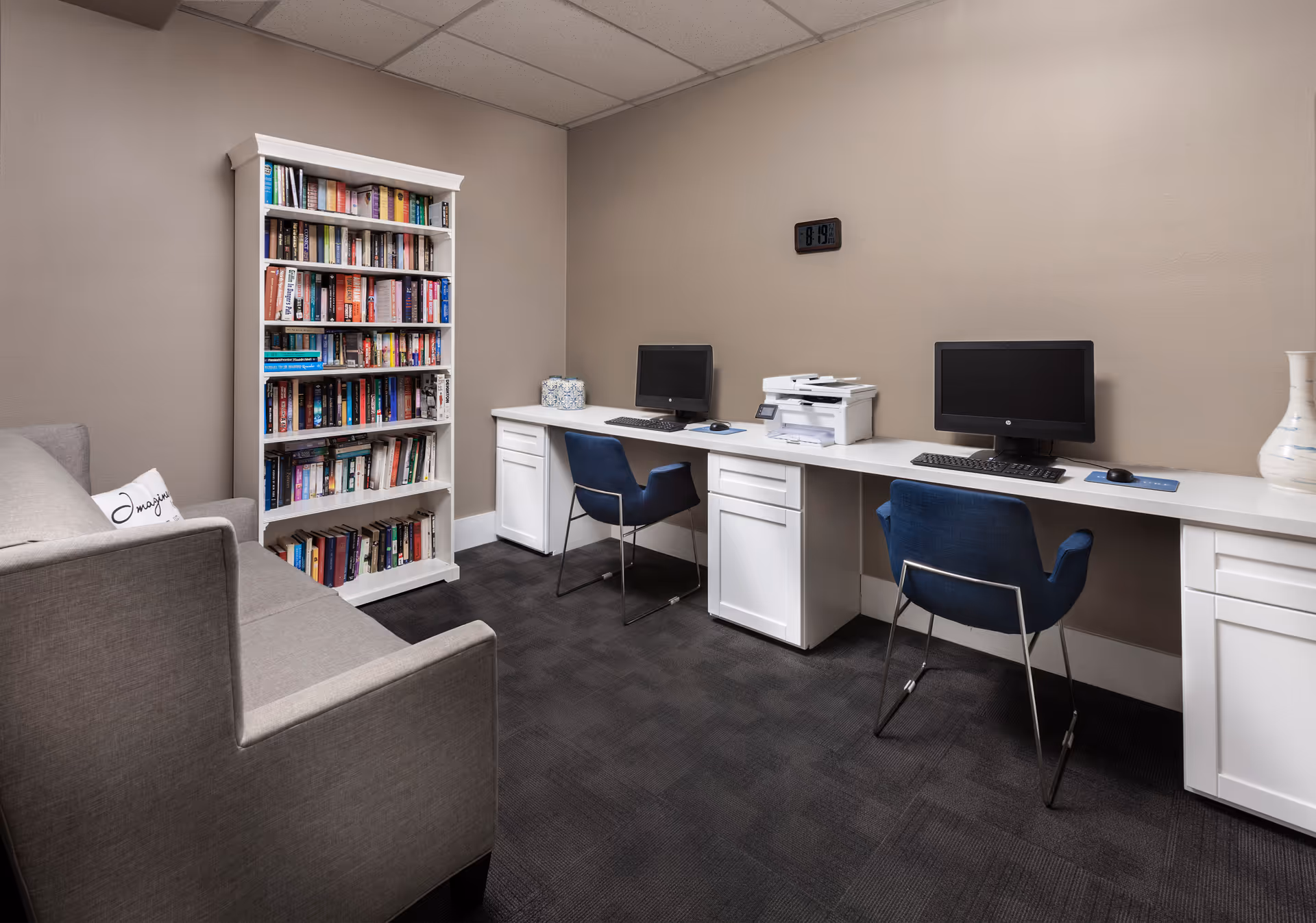 A small office or study room with a gray couch on the left, a white bookshelf filled with books, and a long white desk with two computer workstations and a printer. Two blue chairs are placed at the desk, and a clock on the beige wall shows the time as 8:19.