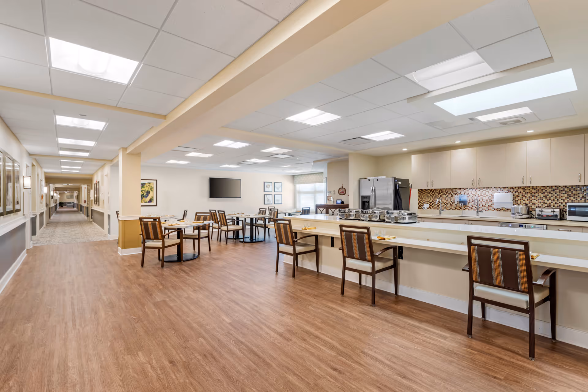 A spacious dining area in a senior living facility with wooden floors, several tables and chairs set for meals, a long counter with chairs, and a kitchen area with cabinets, a refrigerator, and appliances. The room is well-lit with ceiling lights and has a hallway extending to the left.