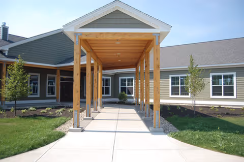 Entrance walkway to a single-story building with a covered wooden pergola structure, surrounded by green grass and small trees under a clear blue sky.