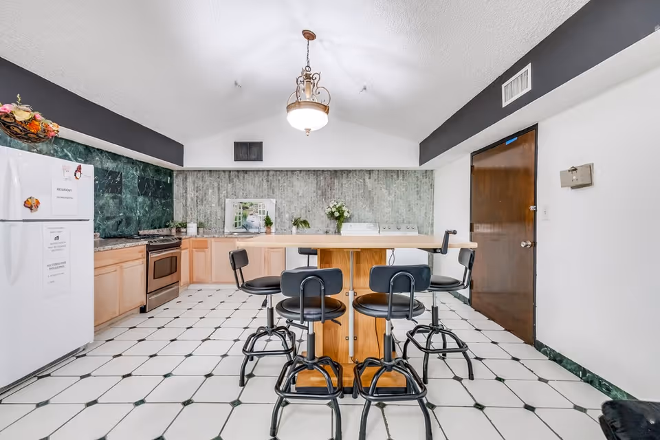 A kitchen area with a white refrigerator on the left, light wood cabinets, a stainless steel stove, and a central island with four black swivel bar stools. The floor is white tile with black accents, and the walls have a green marble-like backsplash and a textured gray wall. A decorative light fixture hangs from the ceiling, and there is a wooden door on the right side.