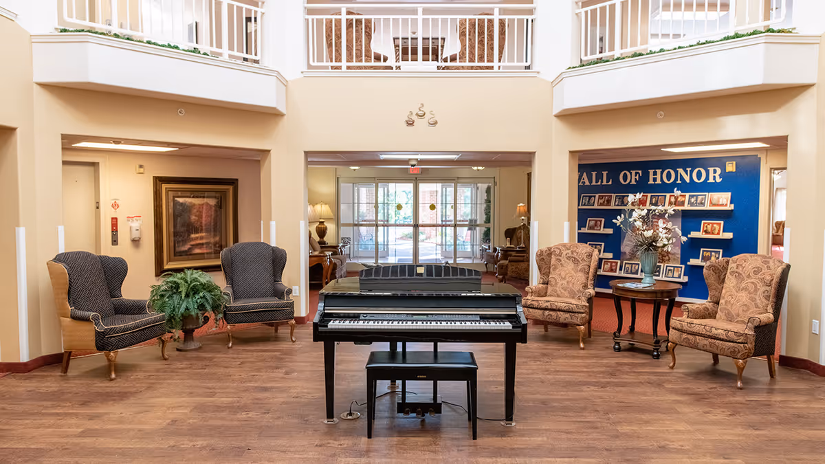 A spacious senior living facility common area with a black piano and matching bench in the center. Surrounding the piano are four upholstered armchairs, two on each side, with a small round table holding a vase of flowers on the right. The back wall features a blue 'Wall of Honor' display with framed photos. The area has wood flooring and beige walls, with a balcony railing visible above.