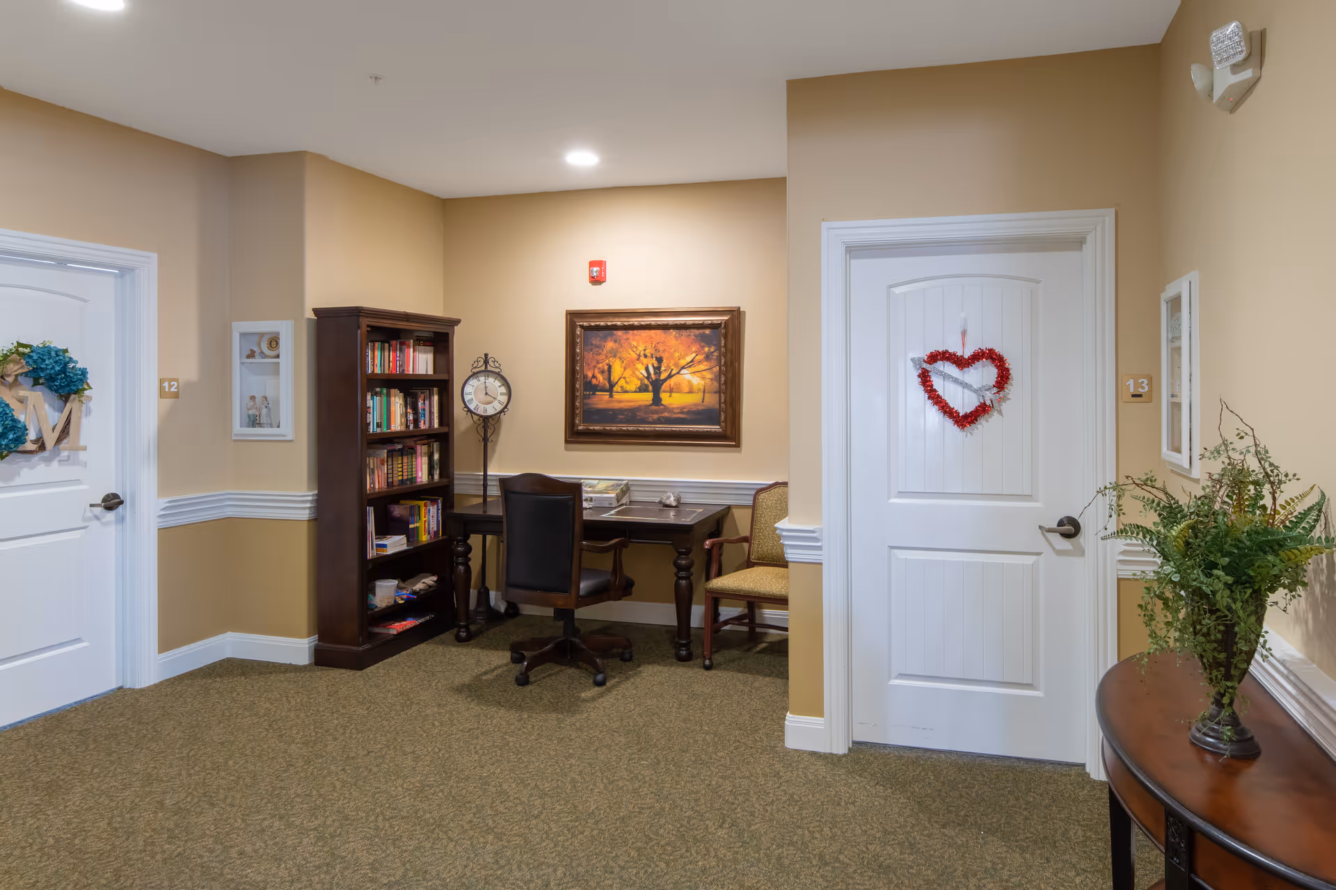 A cozy hallway corner in an assisted living facility with two white doors numbered 12 and 13, decorated with wreaths. Between the doors is a dark wooden bookshelf filled with books, a small desk with a chair, and a framed painting of a tree with autumn leaves on the wall. A round clock stands on the floor next to the bookshelf. On the right side, there is a small wooden table with a green plant in a vase. The walls are painted beige with white trim, and the floor is carpeted.