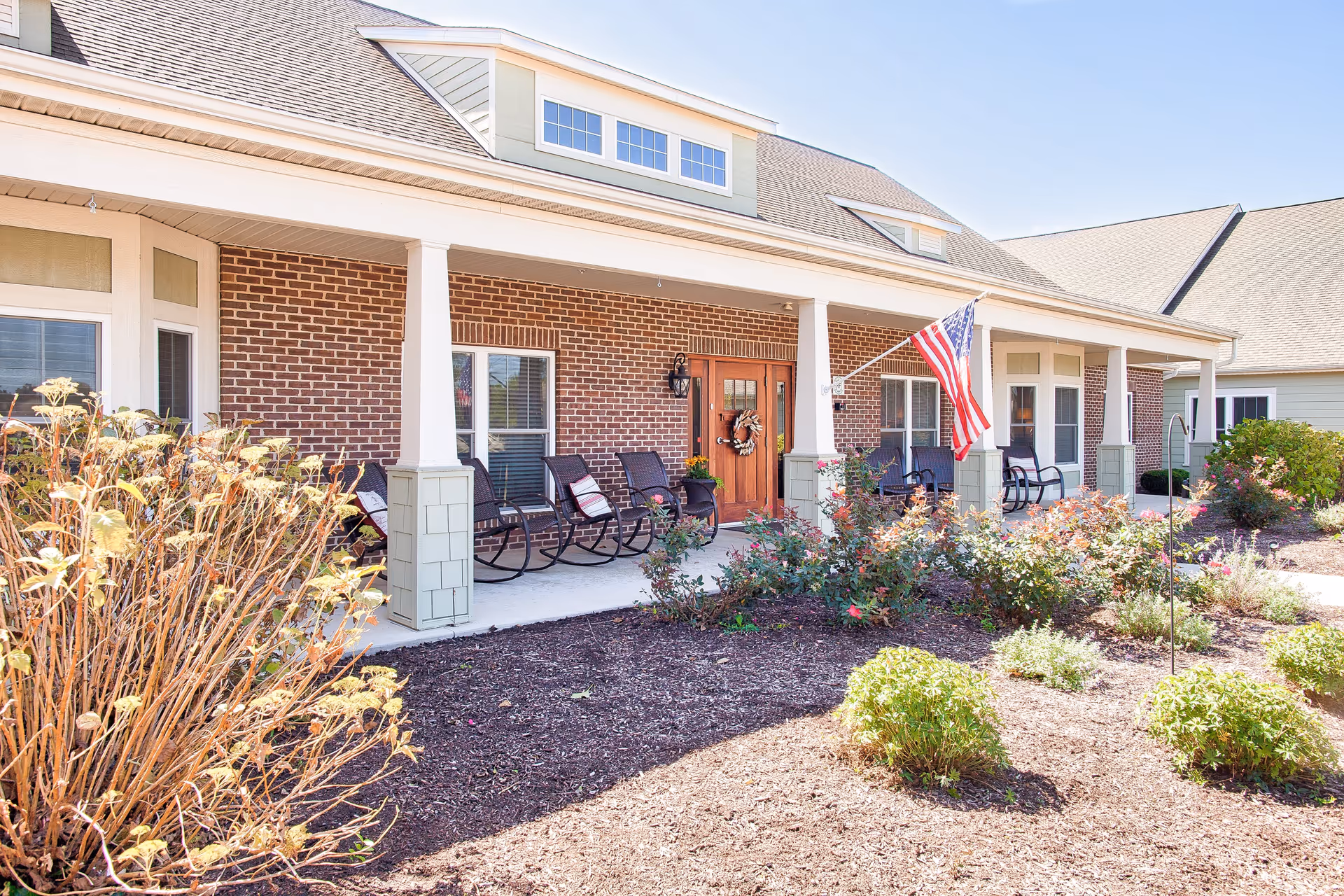 Front porch of a senior living facility with a brick exterior, white columns, several rocking chairs, an American flag, and landscaped garden beds with shrubs and flowers.