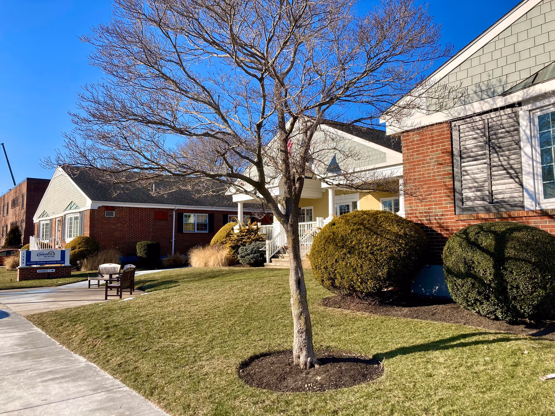 Exterior view of Compass on the Bay Memory Care Assisted Living facility showing a brick building with green siding and white trim. There is a leafless tree in the foreground, neatly trimmed bushes, a grassy lawn, and a sidewalk leading to the entrance. A sign near the sidewalk displays the facility's name.