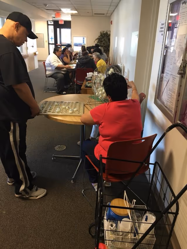 Several elderly people sitting at tables in a hallway playing bingo, with one person standing and another sitting at a small round table operating a bingo cage and board.