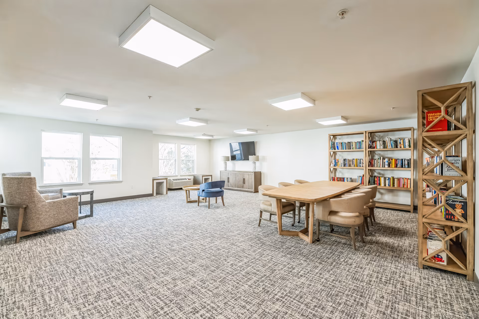 Bright communal lounge with a long wooden table and chairs, bookshelves, a TV cabinet and seating by windows.