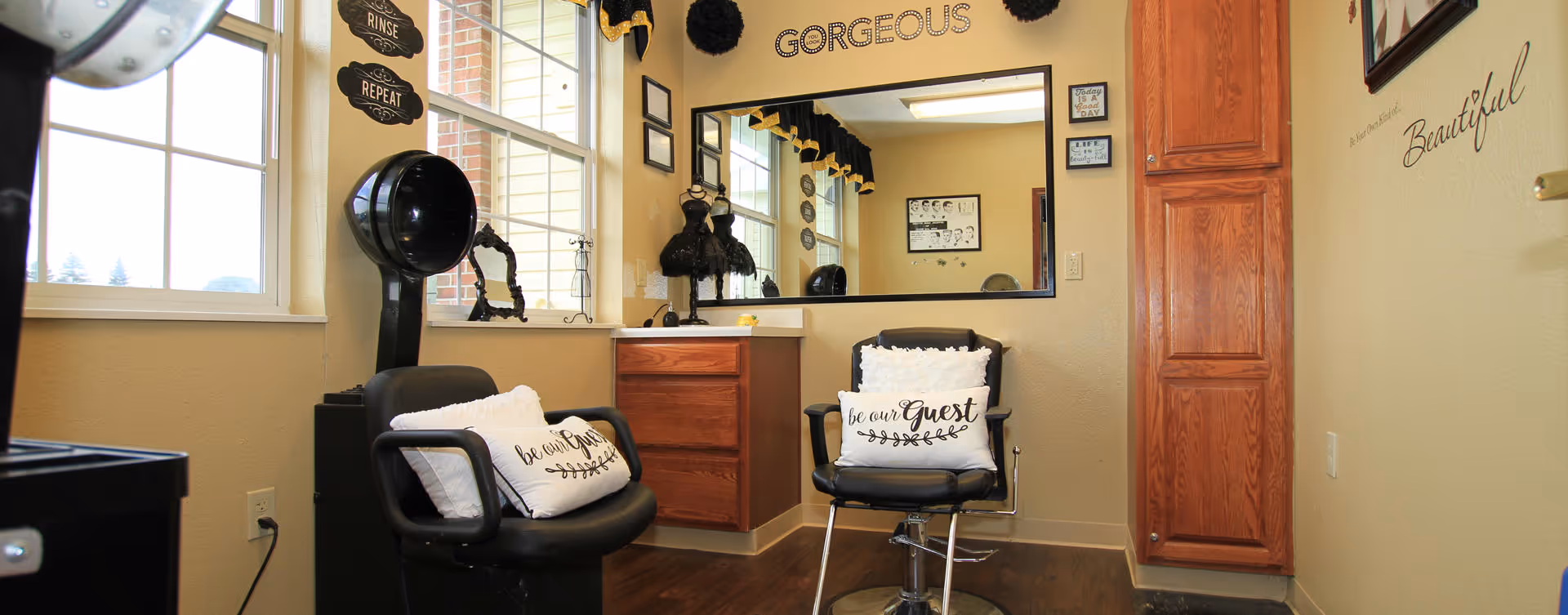 Interior of a salon area in a senior living facility with two black salon chairs, each with a white pillow that says 'be our Guest'. There is a large mirror on the wall with the word 'GORGEOUS' above it, and decorative signs on the walls including 'RINSE', 'REPEAT', and 'Beautiful'. The room has wooden flooring, a window letting in natural light, and wooden cabinetry.