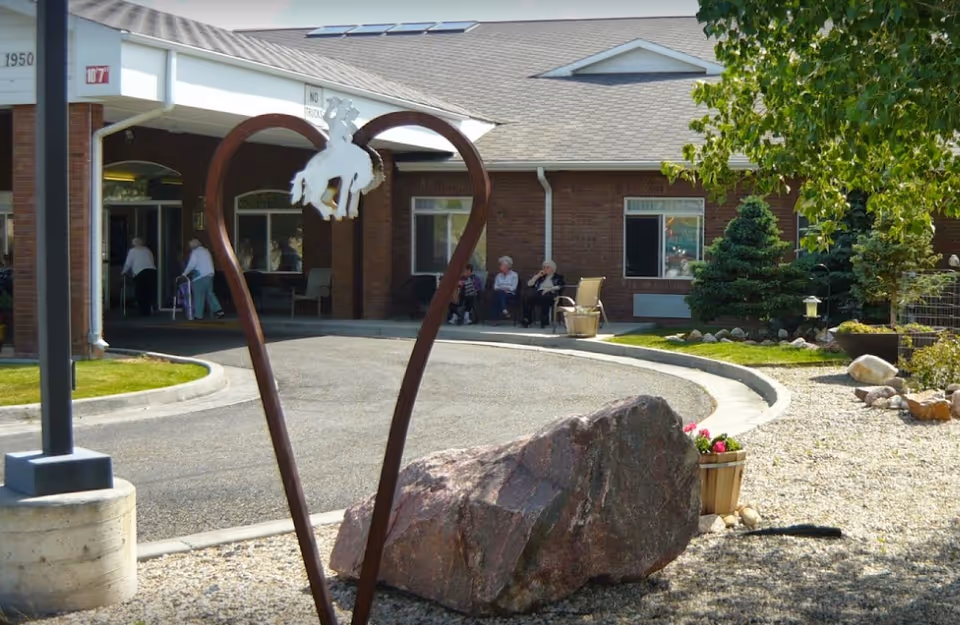 Front entrance of a brick assisted living building with a heart-shaped metal sculpture, a large rock, landscaping, and residents seated near the doorway.