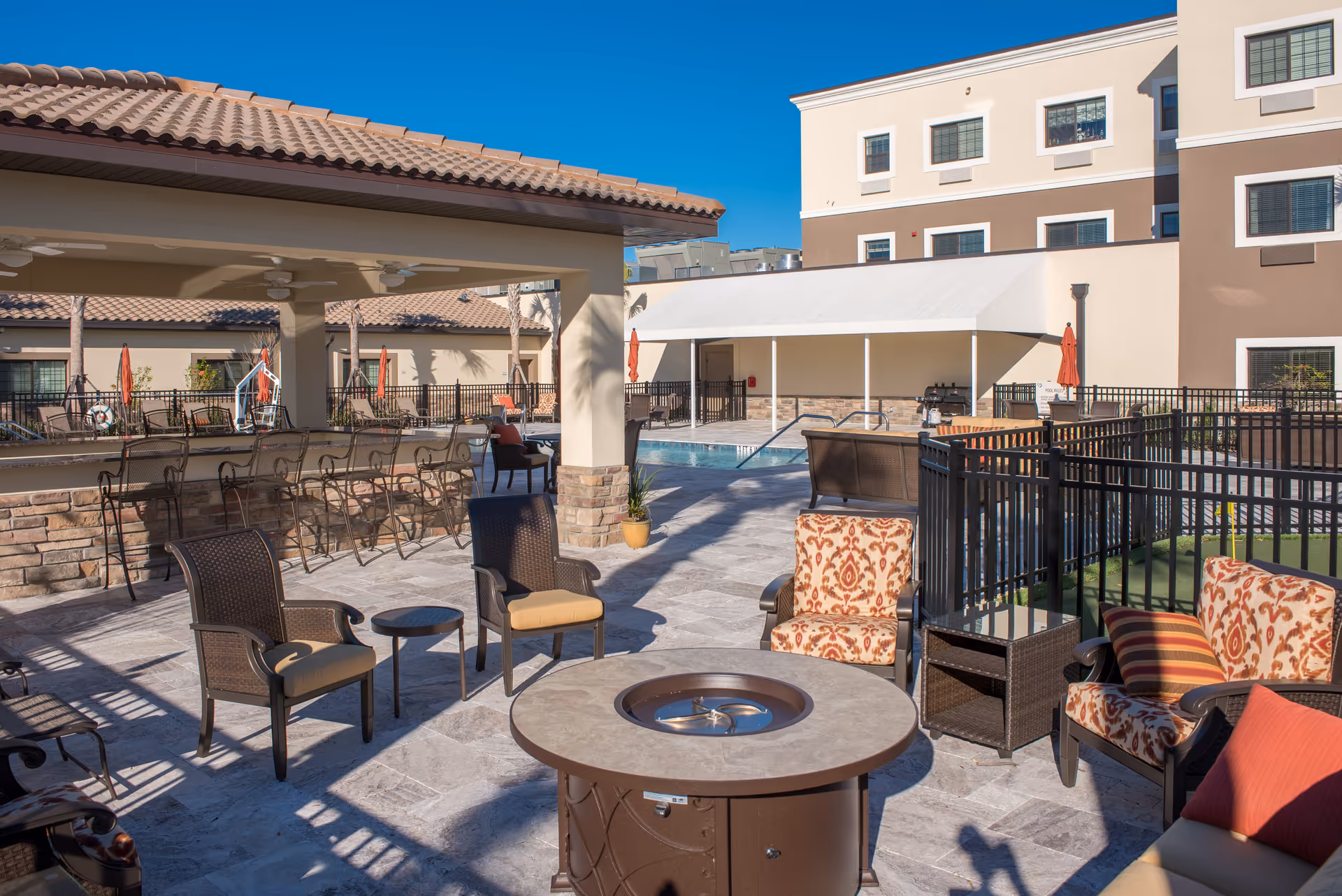 Outdoor patio area at Inspired Living Lakewood Ranch featuring cushioned chairs around a fire pit table, bar seating under a covered area with ceiling fans, and a swimming pool in the background surrounded by fencing and umbrellas.