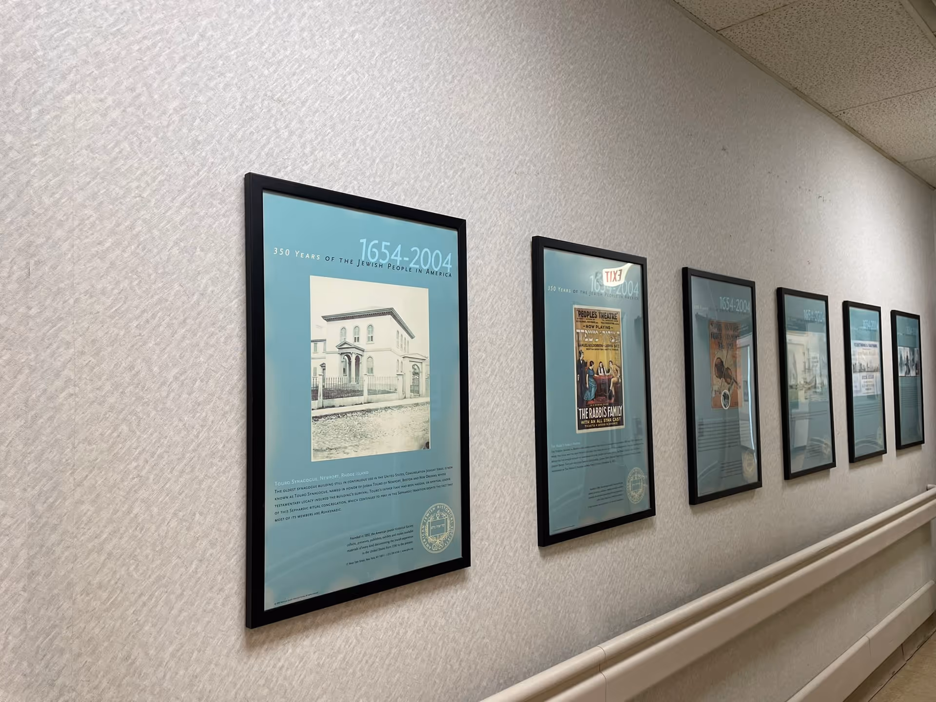 A hallway with six framed posters on the wall, each displaying historical images and text about 350 years of the Jewish people in America from 1654 to 2004. The wall has a light textured wallpaper and a handrail along the length of the hallway.