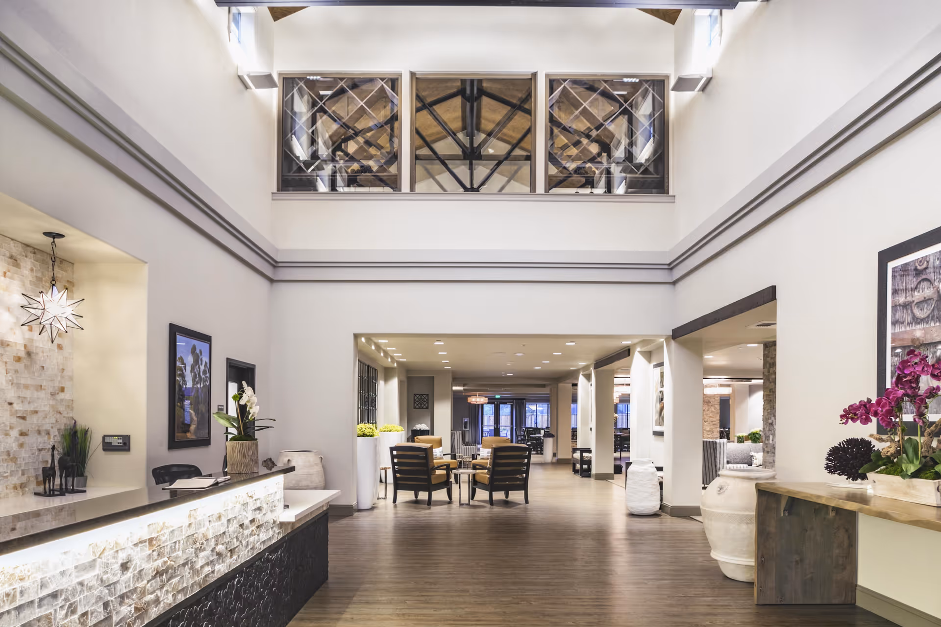 Spacious and well-lit interior lobby area of The Oaks at Nipomo featuring a reception desk with decorative lighting and plants, seating area with chairs and tables, high ceilings with exposed beams visible through upper windows, and tasteful wall art and decor.