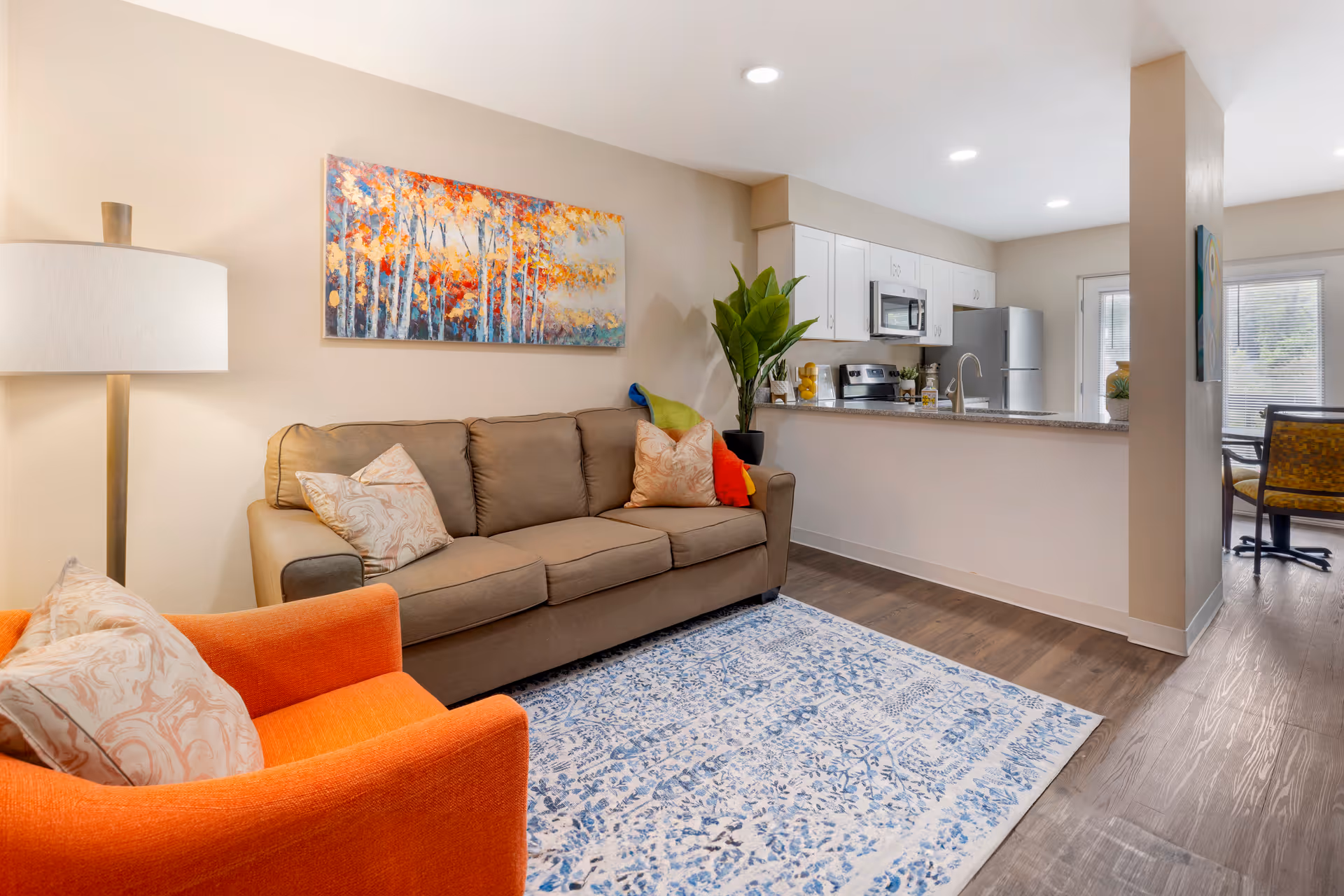 Bright open living room with a sofa, orange armchair, patterned rug and a view into a modern kitchen.