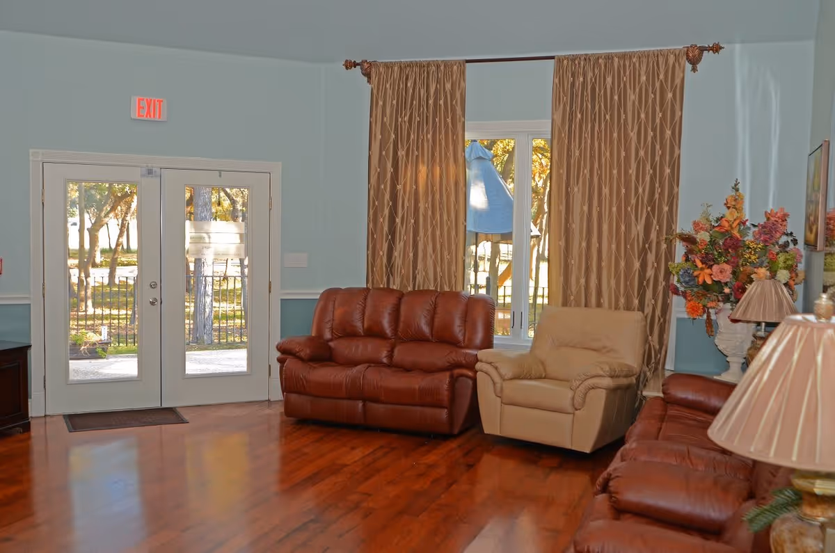 A cozy living room area with two brown leather sofas and one beige armchair arranged on a polished wooden floor. Behind the seating is a large window with brown patterned curtains, and a glass door with an exit sign above it. A floral arrangement in a white vase and a table lamp with a beige shade are visible on the right side.