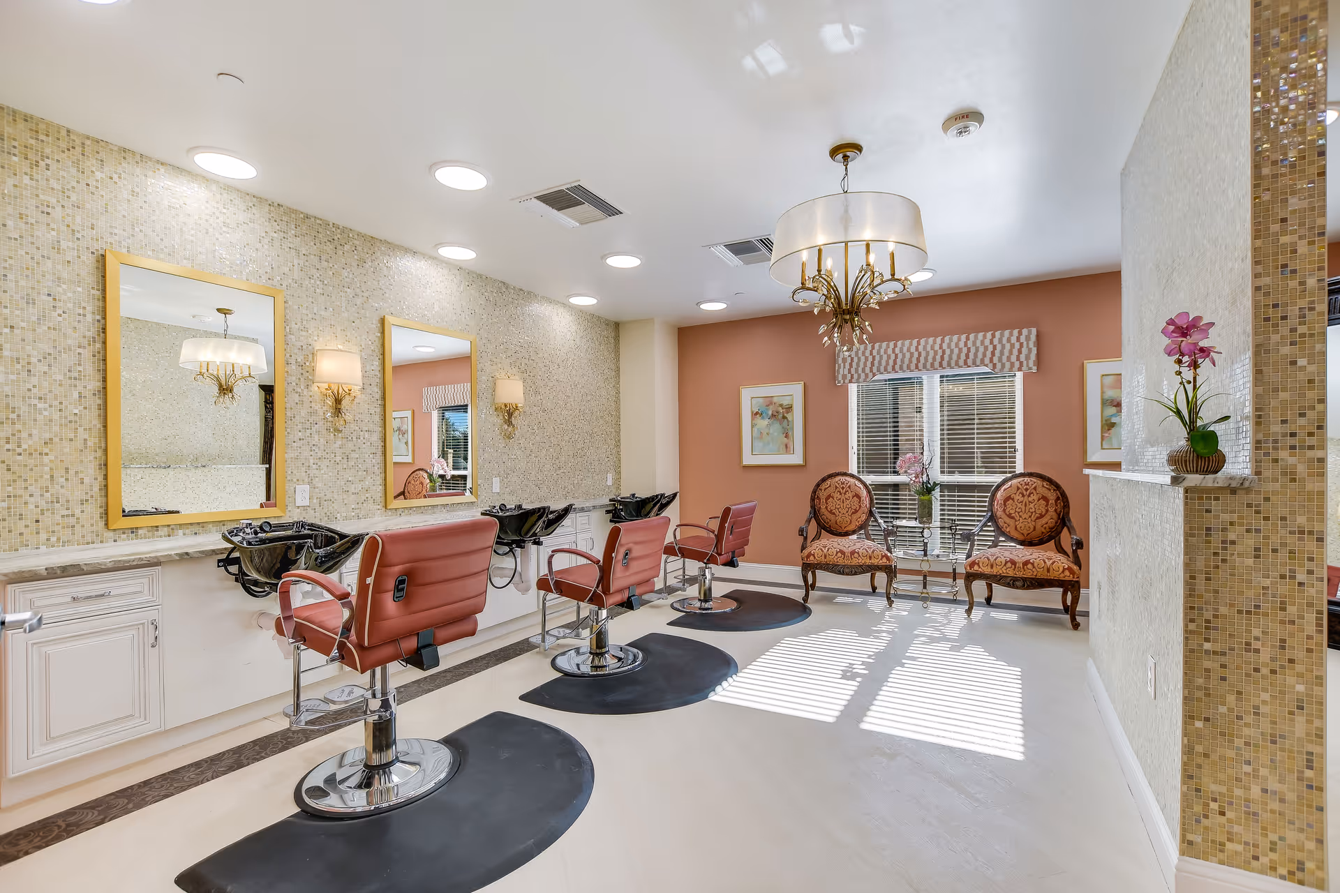 Interior view of a senior living facility's hair salon area featuring three red salon chairs in front of mirrors and sinks. The room has a mosaic tiled wall, soft lighting fixtures, a chandelier, and a seating area with two upholstered chairs and a small table near a window with blinds.