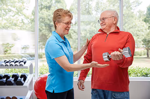 An elderly man in a red long-sleeve shirt lifting a dumbbell while a female fitness instructor in a blue polo shirt encourages him in a bright exercise room with large windows showing greenery outside.