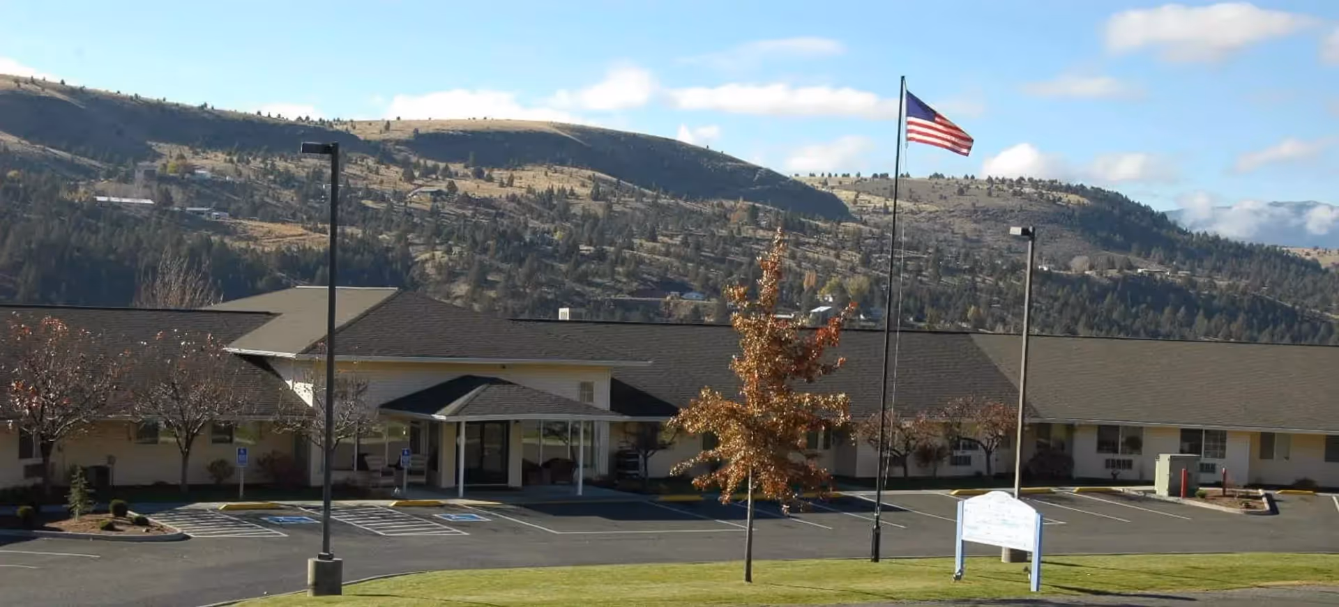 Exterior view of a single-story senior living facility building with a covered entrance, an American flag on a flagpole, a parking lot with marked spaces, and hills in the background under a partly cloudy sky.