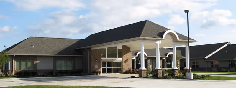 Exterior view of Plum Creek Assisted Living facility showing a single-story building with a covered entrance supported by white columns, brick walls, and a dark shingled roof under a partly cloudy sky.