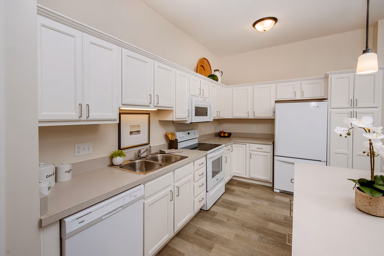 A clean, modern kitchen with white cabinets and beige countertops. The kitchen features a double sink, a white dishwasher, a white electric stove with an overhead microwave, and a white refrigerator. There are decorative items such as a small plant, a framed picture, and a potted orchid on the counter. The floor is light wood, and the ceiling has a flush mount light fixture and a hanging pendant light.