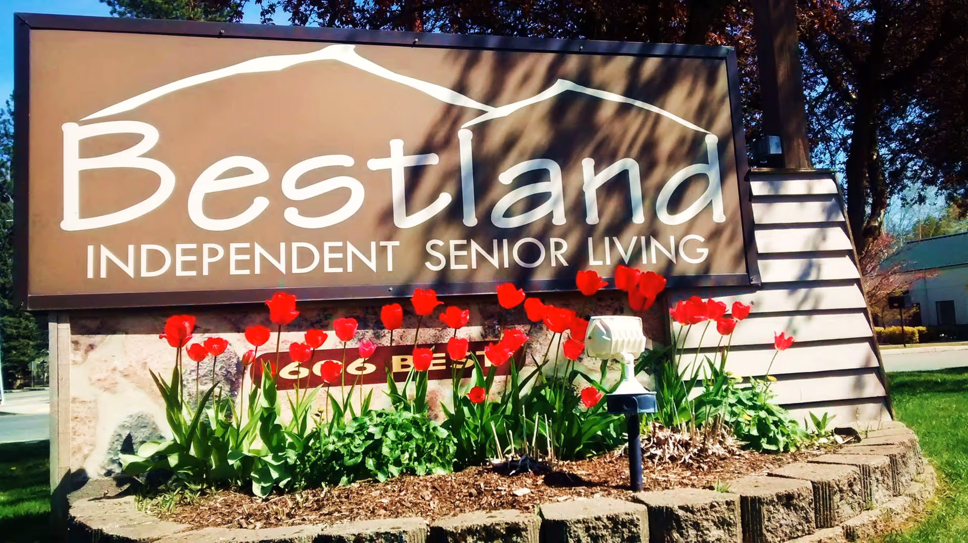 Outdoor sign for Bestland Independent Senior Living with red tulips planted in front and trees in the background on a sunny day.
