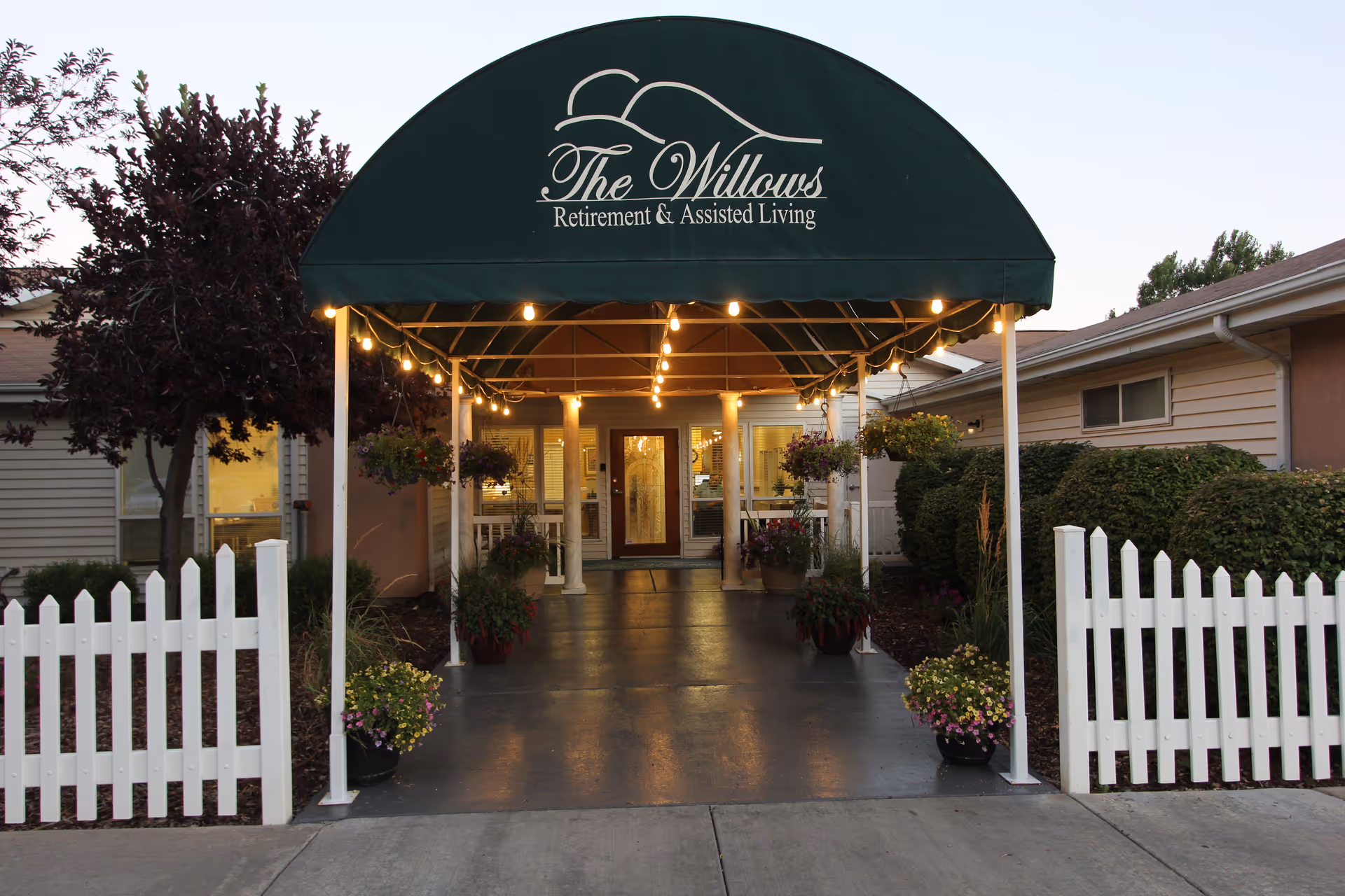 Entrance to The Willows Retirement & Assisted Living facility with a green canopy displaying the facility name. The walkway is lined with potted plants and hanging flower baskets, leading to a glass door entrance. White picket fences and bushes flank the walkway.