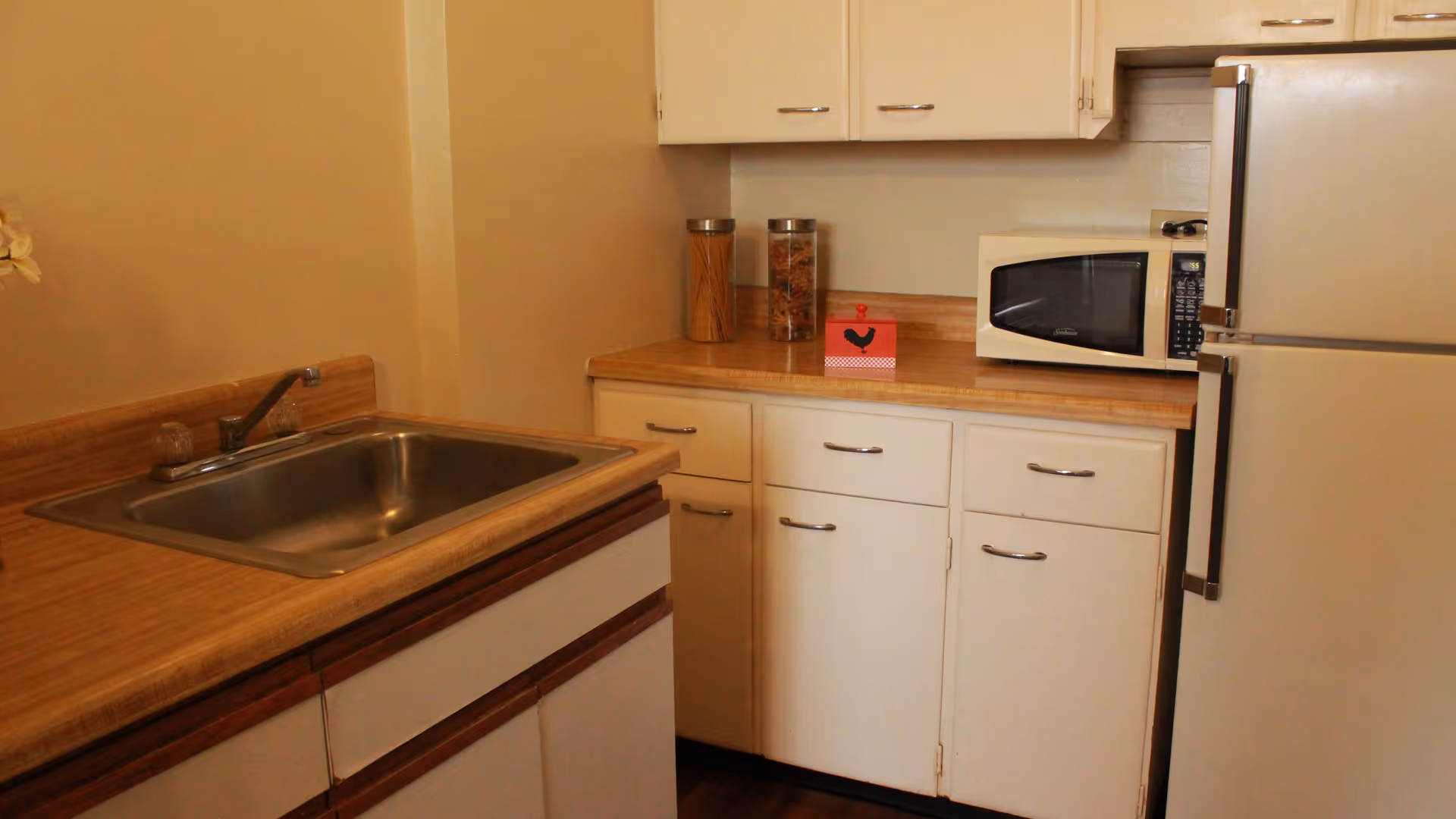 A small kitchen area with a stainless steel sink, wooden countertops, white cabinets, a microwave, a refrigerator, and two jars containing pasta and cereal on the counter.