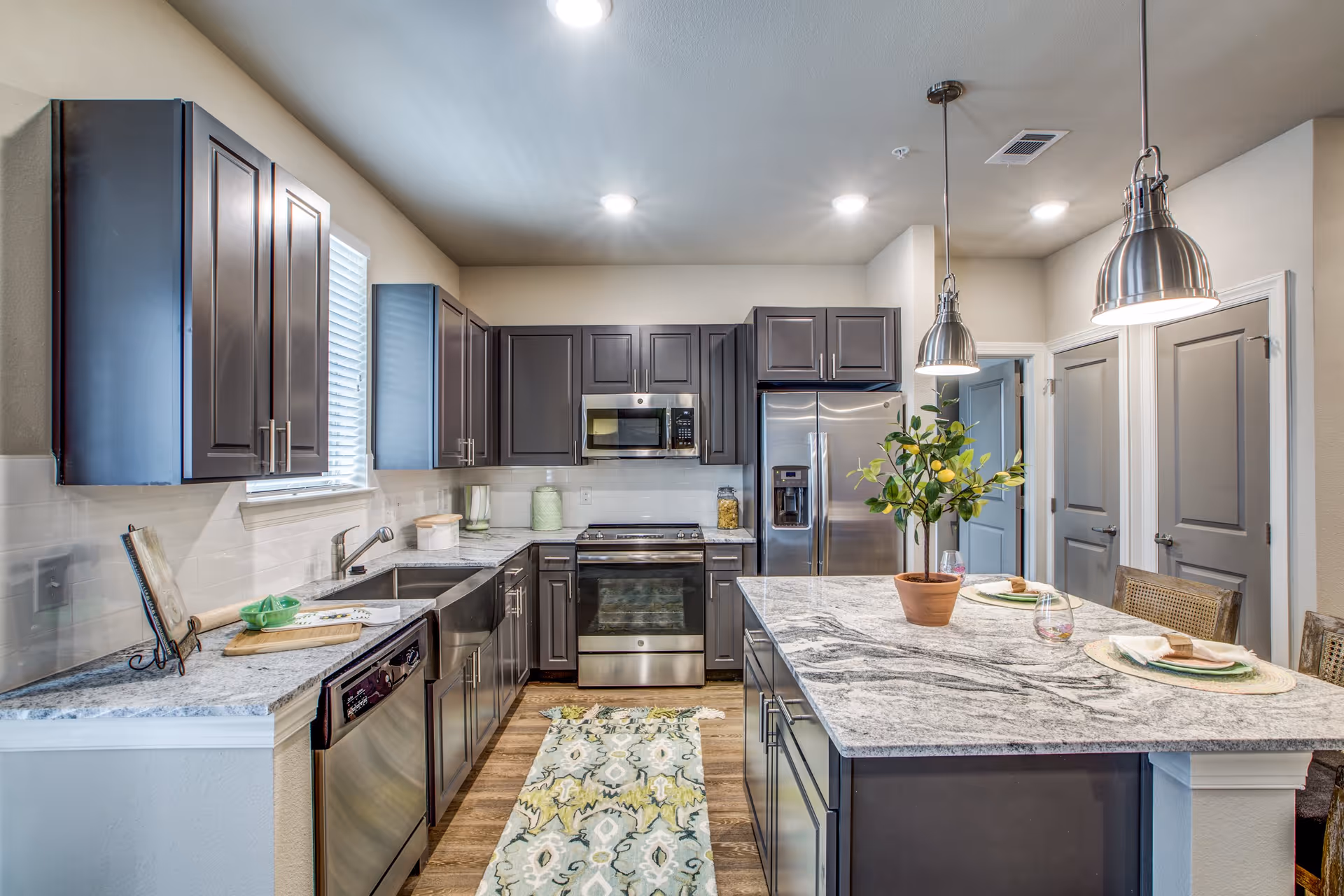 Modern kitchen with dark cabinetry, stainless steel appliances including a refrigerator, oven, microwave, and dishwasher. The kitchen features a large island with a marble countertop, two pendant lights hanging above, and a potted plant centerpiece. There is a patterned rug on the wooden floor and three place settings on the island. The walls are light-colored with two closed doors visible in the background.