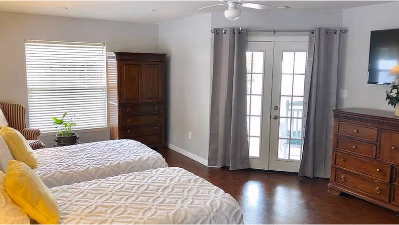 A bright bedroom with two twin beds covered in white textured bedspreads and yellow pillows. There is a wooden dresser and armoire, a window with blinds, a potted plant, and French doors with gray curtains leading outside. A ceiling fan and a wall-mounted TV are also visible.