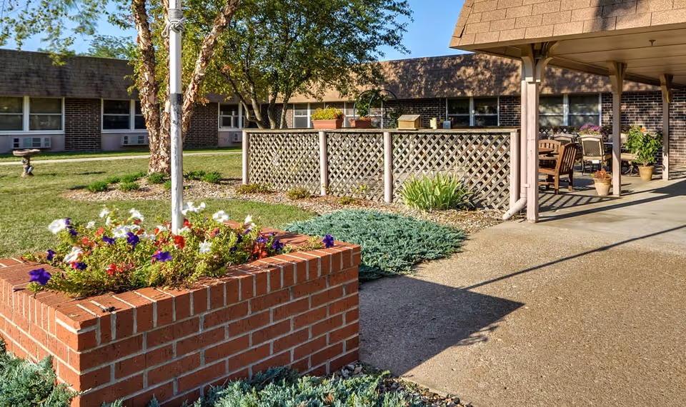 A sunny courtyard with a brick flower planter, lattice fence, and covered patio seating in front of a low brick building.