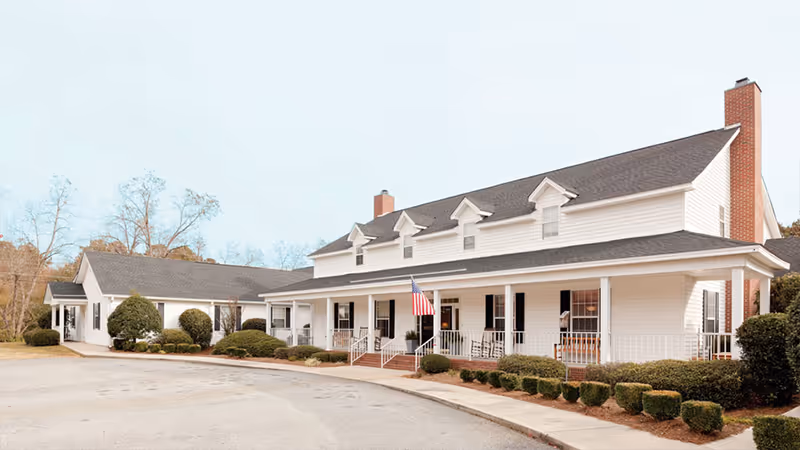 Front view of a two-story white senior living building with a covered porch, American flag, and landscaped shrubs.