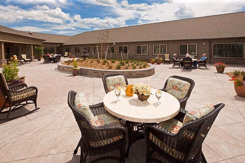 Outdoor courtyard area of Canyon Creek Memory Care Community featuring a round table with four cushioned wicker chairs, a flower centerpiece, and two glasses. The courtyard has a circular raised garden bed with plants and trees, additional seating areas with people sitting and conversing, and a building with multiple windows surrounding the space under a partly cloudy sky.