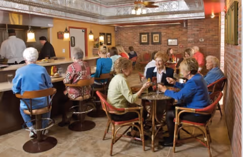 Several elderly residents sit at tables and a counter chatting and dining in a brick-walled communal dining area.
