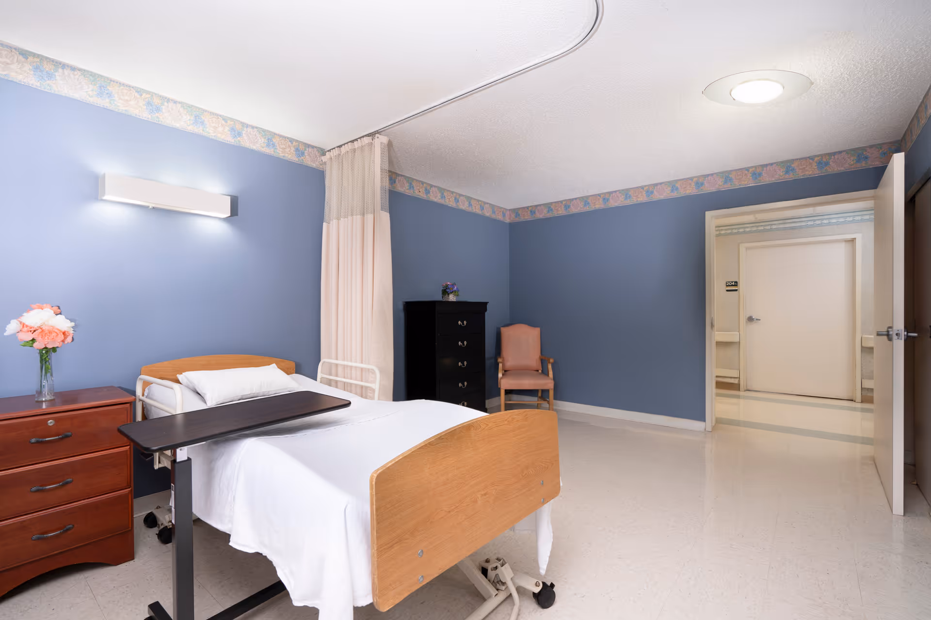 A hospital-style room with a single bed featuring a wooden headboard and footboard, white bedding, and an overbed table. The walls are painted blue with a floral border near the ceiling. There is a wooden nightstand with a vase of pink and white flowers, a black chest of drawers, and a pink upholstered chair. The room has a ceiling light and an open door leading to a hallway with beige walls and doors.