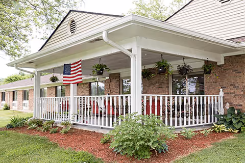 Exterior view of a brick building with a covered porch featuring white railings and hanging plants. An American flag is displayed on the porch, and there are red chairs visible inside. The surrounding area has green grass, shrubs, and mulch beds.