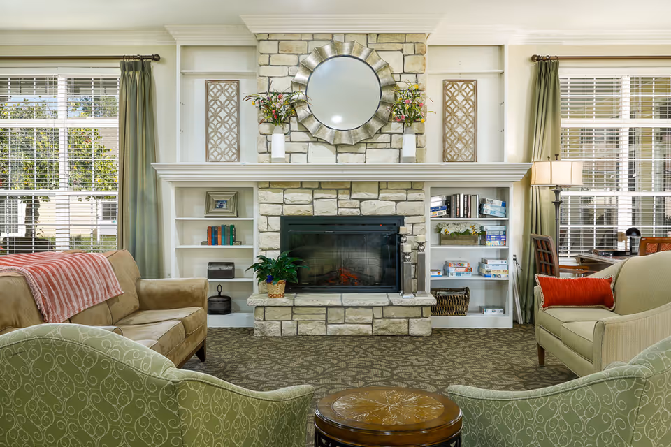 A cozy living room with a stone fireplace in the center, flanked by built-in white bookshelves filled with books and board games. Above the fireplace is a decorative round mirror with a metallic frame and two vases with flowers. The room has large windows with white blinds and green curtains. There are two beige sofas, one with a red throw blanket and the other with a red pillow, and two green patterned armchairs facing a round wooden coffee table. A floor lamp and a small desk with a chair are visible near the right window.