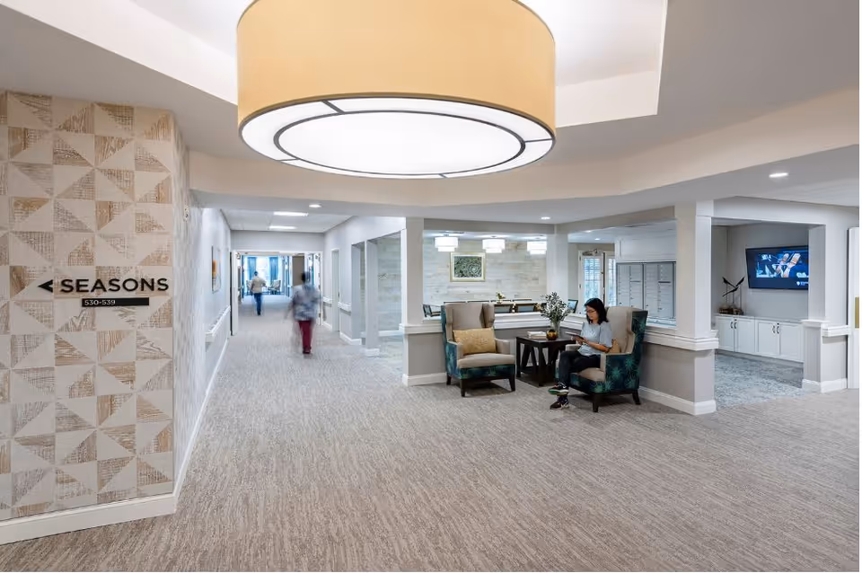 Interior common area of a senior living facility showing a hallway and seating area with armchairs, a large overhead light, a person reading, a TV, and a wall sign reading "SEASONS 530-539".