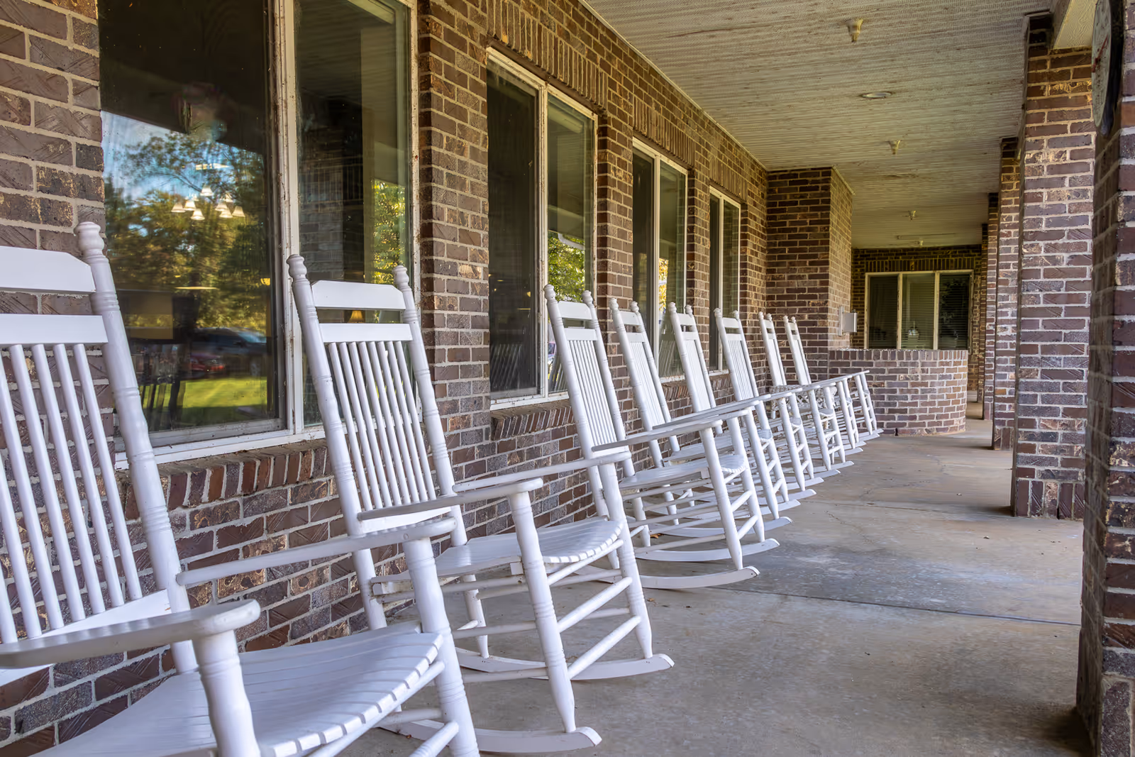 A row of white wooden rocking chairs lined up on a covered brick porch with windows reflecting outdoor greenery.